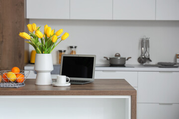 Vase with yellow tulip flowers, modern laptop, fruits and cup of coffee on wooden table in light kitchen