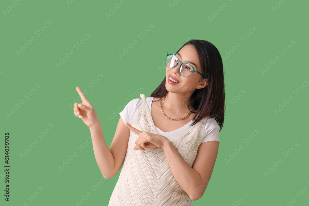 Wall mural Beautiful Asian woman in stylish eyeglasses pointing at something on green background