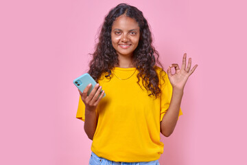 Young casual happy Indian woman millenial with smartphone in hand shows gesture everything is ok to answer question how are you or approve your act stands on plain pink background.