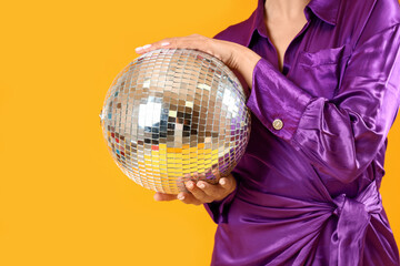 Fashionable young woman in elegant purple dress with disco ball posing on yellow background