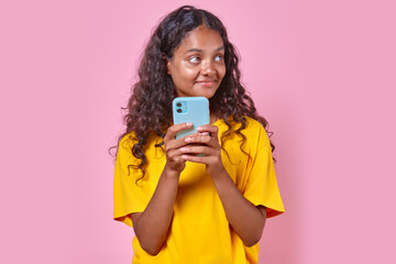 Fototapeta premium Young cute cheerful Indian woman with phone looks away coming up SMS message for boyfriend or comment for blog in social network stands on isolated pink background. Blogger, influencer