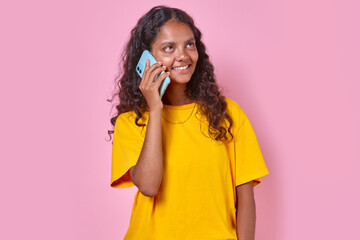 Young positive beautiful Indian woman teen in casual clothes makes phone call to friend from school and smiles broadly discussing plans for weekend or summer vacation stands in pink studio.