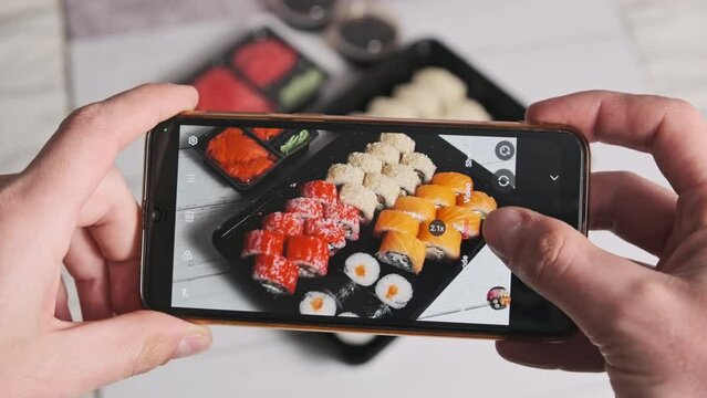 Photographing sushi on a smartphone for social networks. Male hands taking photos of prepared sushi rolls in delivery box on white photo background. Blogger shoots Japanese food on mobile phone camera