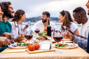 Young people enjoying delicious barbecue dinner party drinking red wine - Multiracial family having diner time together outside - Happy friends eating fresh food sitting at restaurant table