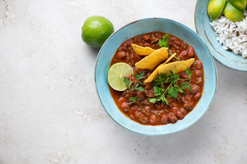 Chili con carne in a turquoise bowl served with nachos chips, mixed rice and lime wedges, top view on a light-beige stone background, horizontal shot, copy space