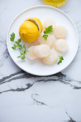 White plate with raw scallops, lemon and fresh parsley, vertical shot on a white marble background with space, high angle view