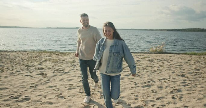 Boy and girl fool around together. Couple in love holding hands and having fun at background of lake and sky. Lovers laughing and running on sand. Male and female satisfied with time spending.
