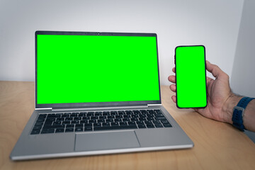 Mockup image of a man holding a mobile phone with green screen and a laptop with blank green screen on table