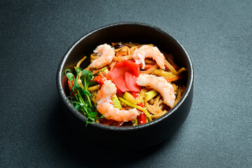 Udon pasta with shrimp, vegetables and ginger. In a black plate. Japanese cuisine. On a dark background, close-up.