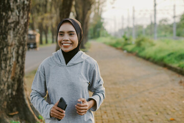 asian muslim woman running and exercising while holding mobile phone outdoors excitedly