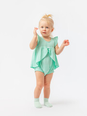 A 2-year-old girl with a questioning expressive look spreads her hands to the side on a white background in a green bodysuit dress.