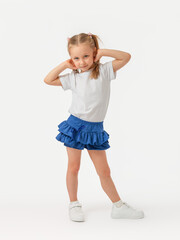 A 5-year-old girl poses with her hands behind her head on a white background and looks expressively at the camera in a white T-shirt, blue skirt and sneakers.