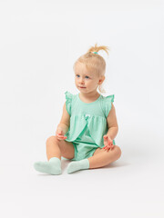 A Caucasian girl of 2 years old sits and attentively looks and studies something on a white background in socks and a green dress. Expressive look.