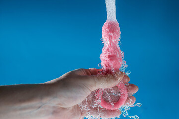 Woman holding pink anal beads under running water on blue background. Sex toy hygiene concept. - obrazy, fototapety, plakaty