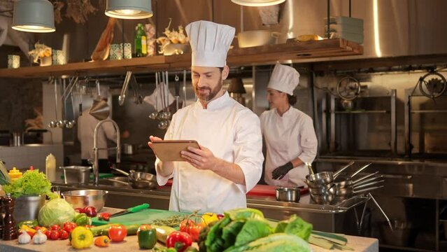 Chef uses digital tablet and keeps list of ingredients needed to prepare dishes. Bearded cook in special uniform is searching for new recipes for tasty and healthy food on Internet.