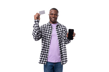 a handsome 30 year old African guy dressed in a plaid black and white shirt demonstrates a bank card mockup and a smartphone in front of him against the background with copy space