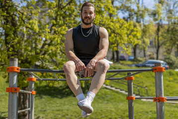 Athletic smiling man in sportswear relaxing after pull ups exercises on horizontal bar. Middle eastern guy on playground. Sports health, fitness routine, workout. Strength and motivation. Outdoor gym