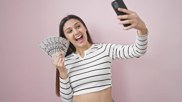 Young beautiful hispanic woman taking selfie holding dollars over isolated pink background