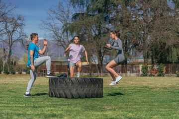 Obraz premium Fit sports friends exercising in the park, on a sunny day. They are doing jumps on a big truck tire