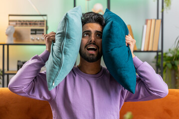 Repair work at neighbours. Irritated indian man relaxing on couch cover ears with pillows annoyed...