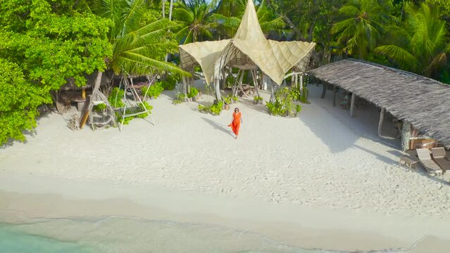 Beautiful sexy girl in a red dress walking down the white sand beach on a paradise tropical island on bamboo bungalows background on Thinadhoo, Maldives. Aerial drone view 4K.