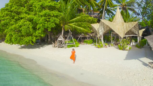 Beautiful sexy girl in a red dress walking down the white sand beach on a paradise tropical island on bamboo bungalows background on Thinadhoo, Maldives. Aerial drone view 4K.