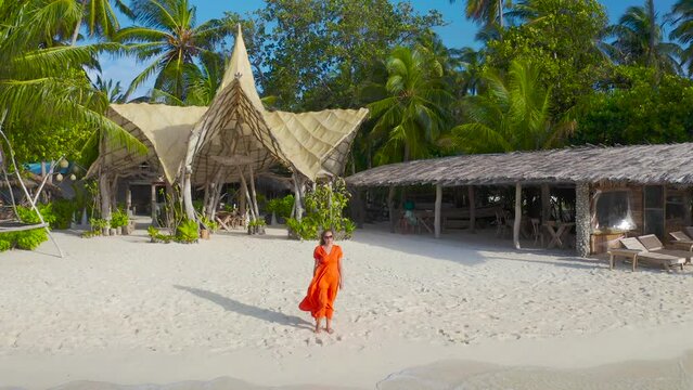 Beautiful sexy girl in a red dress walking down the white sand beach on a paradise tropical island on bamboo bungalows background on Thinadhoo, Maldives. Aerial drone view 4K.