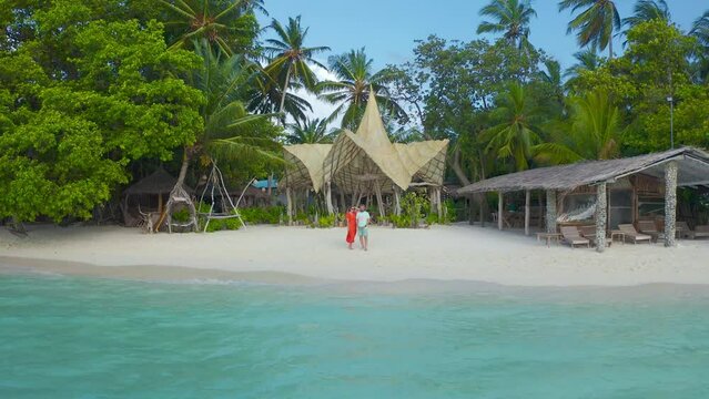 Happy couple, man and woman on red dress the white sand beach on a paradise tropical island on bamboo bungalows background on Thinadhoo, Maldives. Aerial drone view 4K.