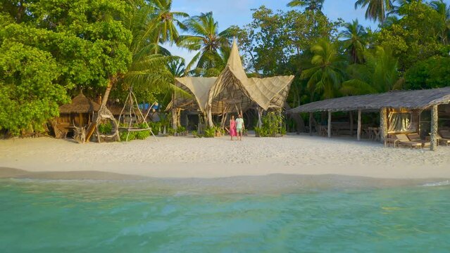 Happy couple, man and woman on red dress the white sand beach on a paradise tropical island on bamboo bungalows background on Thinadhoo, Maldives. Aerial drone view 4K.