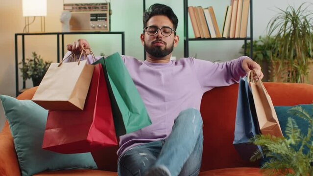 Young indian man happy shopaholic consumer came back home after shopping sale with bags. Portrait of hindu guy satisfied received parcels purchase from online order at home apartment room on sofa