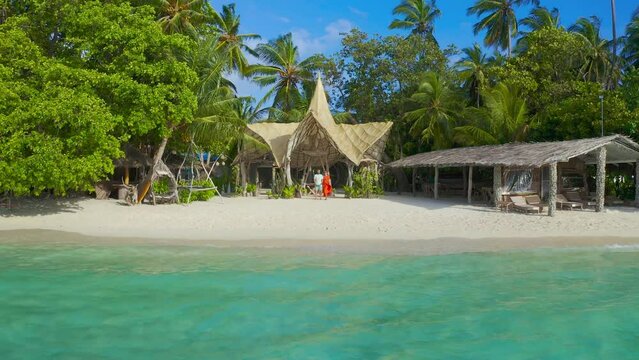 Happy couple, man and woman on red dress the white sand beach on a paradise tropical island on bamboo bungalows background on Thinadhoo, Maldives. Aerial drone view 4K.