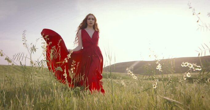 Woman model in fashionable red dress walks across field at sunset like fashion queen. Elegant gait and graceful figure of woman. Queen of style elegance in impeccable dress. Incredible fashion look.