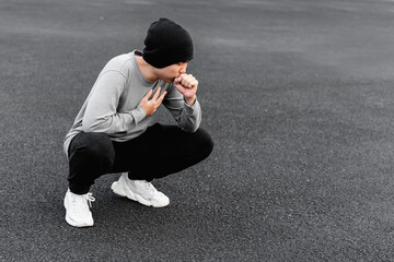 Asian man is coughing and hand covering his chest, half squatting outdoors. 