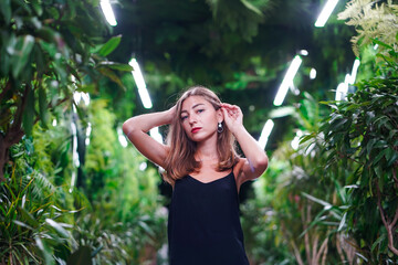Young woman wearing black dress on thin straps stands among green plants inside orangery. Fashionable girl posing in greenhouse or botanical garden. Young female model in evening outfit