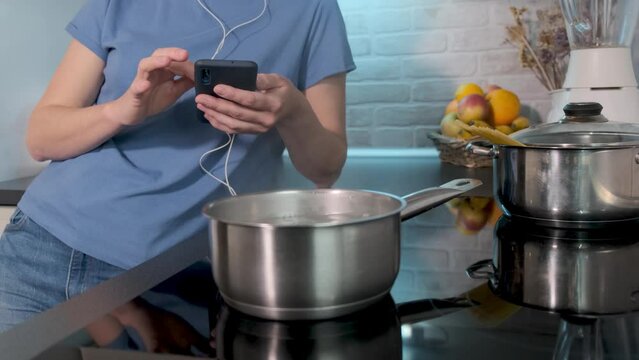 Young caucasian woman watch in mobile phone, social media, stories. Boiling water in saucepan on black induction cooking. Modern kitchen, household appliances, cookware. Girl use gadget, search recipe