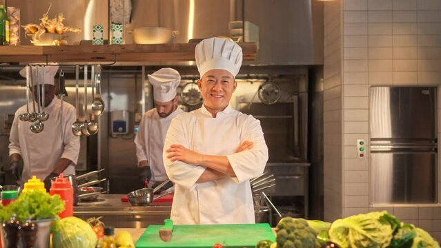 Asian male cook stands with crossed arms, looks at camera and smiling. Satisfied chef at workplace on background of restaurant kitchen. Professional team of multicultural cooks.