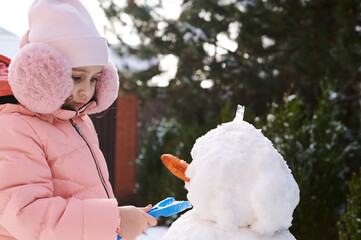 Close-up portrait of a lovely little child girl 5 years old, in pink down jacket and fluffy earmuffs, using shovel building a snowman in the snowy backyard. Leisure activities and games at wintertime