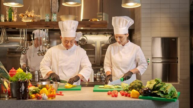 Male and female cooks cooking together at workplace on background of restaurant professional kitchen. Asian and european chefs cutting vegetables and prepearing vegetarian dishes.