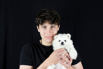 A teenage boy gently holds his little white puppy dog and smiles on dark background, he has long dreamed of such a friend with whom you can walk and talk