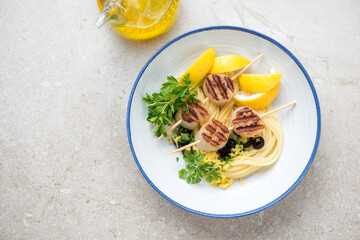 Plate with bbq scallops on skewers, spaghetti, lemon wedges and parsley, horizontal shot on a beige stone background, high angle view, copy space