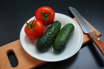 cucumbers and tomatoes on a cutting board with a knife, fresh vegetables on a white plate on a black table, kitchen, cooking, food