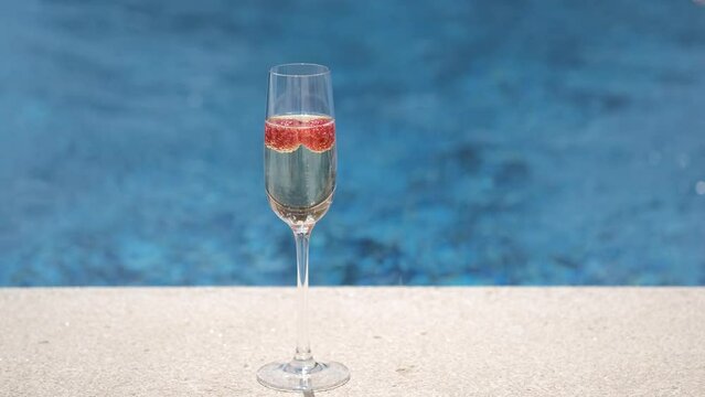 Close up of two glasses with champagne or prosecco with raspberry on swimming pool, sea background. Happy woman takes one glass for drinking cold champagne.  Sparkling white wine on poolside backdrop.