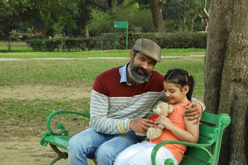 Happy Indian Grand parents with grand daughter in a lush green serene park. They are playing with the ball on park bench.