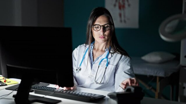 Young beautiful hispanic woman doctor using computer talking on telephone at clinic