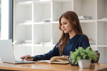 Smiling asian undergraduate teen girl student study in library with laptop books doing online research for coursework, making notes for essay homework assignment, online education e-learning concept