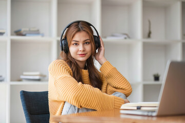 Cute Asian woman in headphones listening to music happily on her laptop enjoying listening to music during work during a relaxing break from studying online.