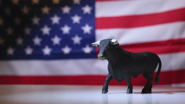 Close-up of a bull figurine symbolizing a bullish market trend, with an out-of-focus American flag waving in the background. This image represents the rise of the stock and cryptocurrency markets