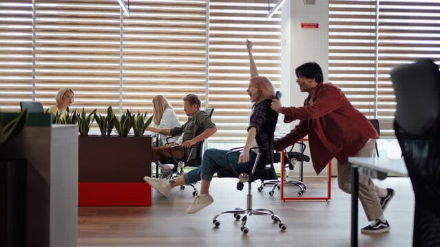 Young Asian man rolls his office co-worker in office chair along the hallway of the office interior