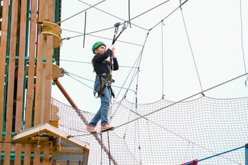 Teenage teen girl in climbing harness equipment, green sports safety helmet. Rope amusement park....