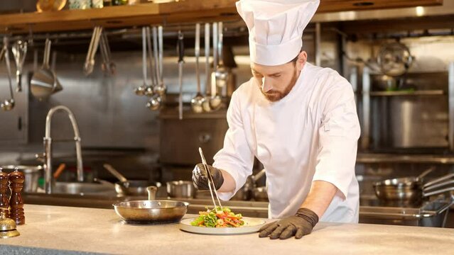 Portrait of professional bearded chef prepares vegetable salad and puts dish on white plate. Male chef dressed in special uniform cooking food in modern restaurant kitchen.